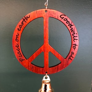May include: A wooden peace sign ornament with the words "peace on earth" and "goodwill to all" engraved on it. A small gold-colored bell hangs from the bottom. The ornament is suspended by a thin wire.