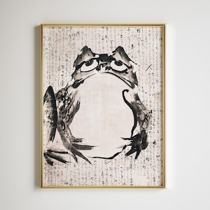 Può includere: Un'opera d'arte incorniciata con un'illustrazione a inchiostro nero e bianco di una rana. La rana ha grandi occhi ed è circondata da calligrafia giapponese. L'opera d'arte è in una cornice dorata.