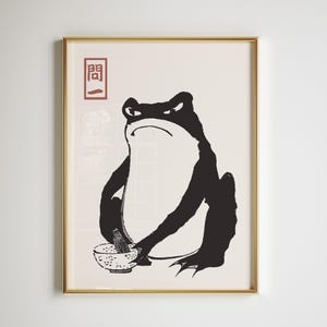 May include: A framed art print featuring a black and white illustration of a frog. The frog is depicted with an angry expression, holding a bowl and whisk. The print includes Japanese text in a red box in the upper left corner. The frame is gold.