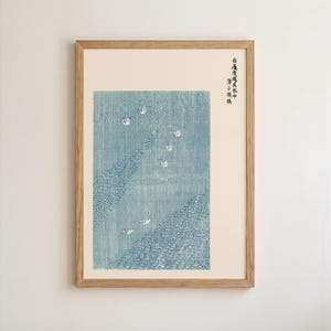 May include: Framed Japanese woodblock print featuring a blue and white seascape with stylized waves and small boats. The artwork is in a light wood frame, adding a touch of natural warmth to the piece. The print has Japanese characters in the upper right corner.