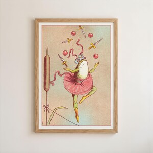 May include: A framed art print featuring a whimsical illustration of a frog in a pink tutu juggling swords and red balls. The frog is adorned with a floral headpiece. A cattail and grass are on the left side of the image. The artwork has a vintage aesthetic.