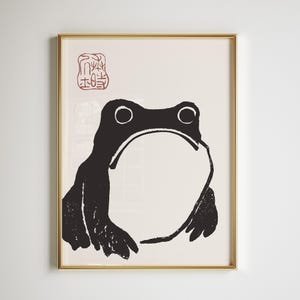 Op de afbeelding: Een ingelijste kunstdruk met een zwart-wit illustratie van een kikker. De kikker heeft grote ogen en een brede mond, met een rode stempel in de linkerbovenhoek. Het kunstwerk staat tegen een crèmekleurige achtergrond en is goudkleurig omlijst.