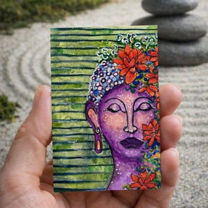 May include: A small, rectangular watercolor painting of a serene Buddha face. The Buddha's face is purple with closed eyes, adorned with red flowers and a blue and white floral crown. The background features green horizontal stripes.