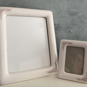 May include: Two off-white ceramic picture frames. The larger frame is rectangular, and the smaller frame is also rectangular. Both frames have a floral design in the corners. The frames are on a white surface with a blue-gray background.