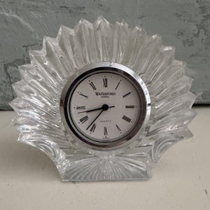 May include: Clear crystal quartz clock with a silver-tone bezel and a white face. The clock is marked "Waterford Crystal" and "Quartz". The clock is in a fan-shaped crystal base.