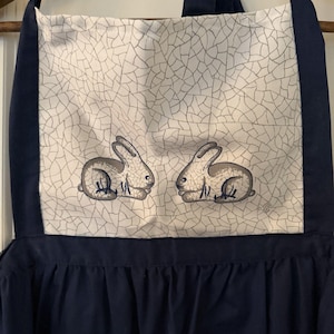 May include: A navy blue apron with a white panel featuring two gray rabbit illustrations. The panel has a cracked pattern design. The apron has a ruffled bottom.