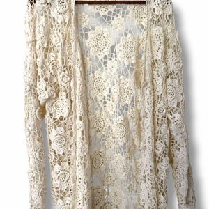 May include: An off-white, long-sleeved cardigan made of intricate crochet lace featuring a repeating floral motif throughout the garment. The open-front design includes a series of small, matching crochet buttons along the right side edge.