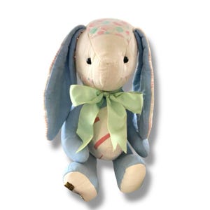 May include: A handmade stuffed bunny with a light blue body and ears, a cream-colored face, and a green bow. The bunny's head is patterned with pastel colors. The bunny is a soft toy.