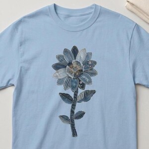 Könnte beinhalten: Hellblaues T-Shirt mit einem Jeansblumen-Design. Die Blume hat blaue Jeansstoff-Blütenblätter in verschiedenen Schattierungen, eine detaillierte Mitte und einen Jeans-Stiel mit Blättern. Das Shirt ist aus weichem Material.