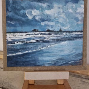 May include: An original oil painting of a seascape, depicting a stormy ocean under a cloudy sky. The artwork features shades of blue and white, with visible brushstrokes. The painting is displayed on a small wooden easel.
