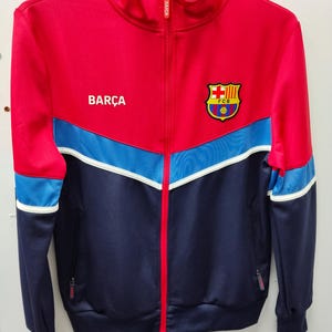 Könnte beinhalten: Eine rot, blau und marineblaue Trainingsjacke mit durchgehendem Reißverschluss und dem Aufdruck "BARÇA" auf der Brust. Die Jacke hat einen Teamlogo-Aufnäher und weiße Akzentstreifen. Sie hat einen Stehkragen und zwei Seitentaschen.