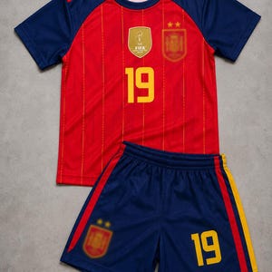 Puede incluir: Un conjunto de fútbol rojo y azul marino que incluye camiseta y pantalón corto. La camiseta presenta finas rayas verticales amarillas, el número 19 en amarillo, un emblema dorado de la FIFA 2010 y un escudo. El pantalón azul marino lleva el número 19.