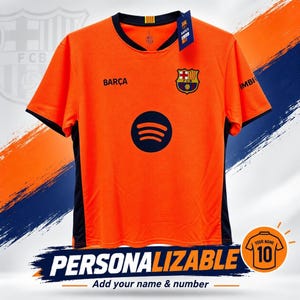 Könnte beinhalten: Ein orangefarbenes kurzärmeliges Sporttrikot mit dunkelblauem Rundhalsausschnitt, dunkelblauen Seitenteilen und dem FC Barcelona-Wappen auf der Brust. Auf der Vorderseite befinden sich das Wort BARÇA und ein dunkelblaues Spotify-Logo. Ein Etikett ist am Kragen befestigt.