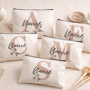 Puede incluir: Bolsas de cosméticos color crema con cremallera negra, cada una con una letra grande y la palabra "Name" en cursiva. Los detalles florales y la paleta de colores neutros crean una estética elegante. También hay accesorios como brochas de maquillaje y una vela.