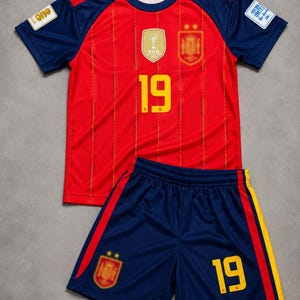 May include: A red soccer jersey with thin yellow vertical stripes, navy blue sleeves, and the number 19 in yellow. It features a FIFA World Cup patch, a team crest, and a Football Unites patch. Matching navy blue shorts include the number 19.