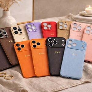 Puede incluir: Una colección de fundas personalizadas para teléfonos en varios colores, incluyendo marrón, naranja, rojo y azul. Cada funda presenta un monograma único en letras doradas. Las fundas están dispuestas sobre una superficie de tonos neutros, con una vela y objetos decorativos en el fondo.