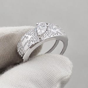 May include: A silver-toned ring featuring a wide band encrusted with numerous small, clear, round-cut stones. A single, prominent pear-shaped clear stone is set at the center of the band, elevated above the surrounding pavé-style stone setting.