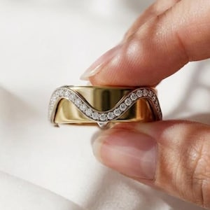 May include: A wide gold band ring featuring a prominent, raised wave-shaped inlay across the center. This decorative wave section is encrusted with a single row of small, clear, round-cut stones that follow the undulating curve of the design.