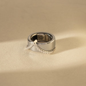 May include: A polished silver-toned ring featuring a wide band with a smooth finish and a V-shaped overlay encrusted with small clear stones. The interior of the band displays a small square QR code and engraved text, visible through the open center.