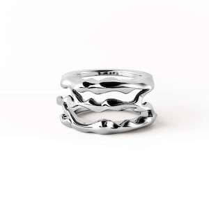 May include: A silver-toned ring featuring a multi-layered design with an irregular, molten-like texture. The band consists of two distinct, wavy, and organic-shaped metal segments that are connected, creating a stacked appearance with a polished finish.