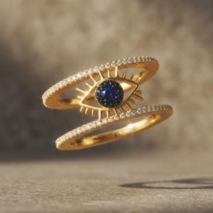 Könnte beinhalten: Ein goldener Doppelring mit einem augenförmigen Mittelstück. Das Auge hat einen dunkelblauen Stein in der Mitte, umgeben von kleinen schwarzen Steinen und goldenen Strahlen. Beide Ringbänder sind mit winzigen klaren Steinen besetzt.