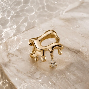 May include: A gold-toned ring featuring an irregular, molten-like metal structure with a smooth, reflective surface. A single, clear, pear-shaped gemstone charm hangs from the bottom of the ring, catching the light with its faceted cut.