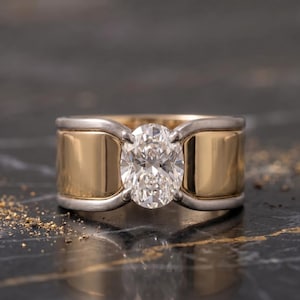 May include: A wide band ring featuring a central oval-cut clear gemstone held by four prongs. The ring is constructed with a polished yellow gold center section flanked by brushed silver-toned metal edges that curve around the central stone setting.