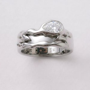 May include: A silver-toned smart ring featuring an organic, fluid metal design with a pear-shaped clear gemstone set on top. The interior band displays the text OURA, the number 11, a small square sensor component, and a QR-style identification code.