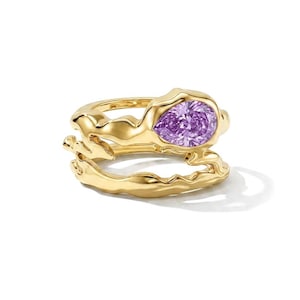 May include: A gold ring featuring a double-band design with an organic, molten-like texture. The upper band is set with a single pear-shaped, faceted purple gemstone. The metal has a polished, reflective finish throughout the entire structure.