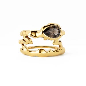 Könnte beinhalten: Ein goldener Ring mit einem organisch geformten, geschmolzen wirkenden Band in offenem, mehrschichtigem Design. Ein einzelner birnenförmiger, durchscheinender rauchbrauner Edelstein ist im oberen Teil des strukturierten, polierten Goldrahmens gefasst.