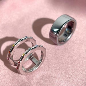 May include: Two silver-toned rings featuring small, colorful embedded gemstones along the outer band, positioned next to a single, wider, plain polished silver-toned band. The rings have a smooth, reflective metallic finish and circular shapes.
