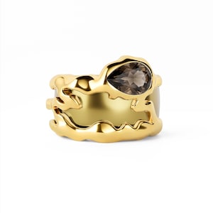 May include: A gold ring featuring an irregular, organic, molten-style band with a smooth, polished finish. A single, teardrop-shaped, translucent brown gemstone is set into the upper edge of the band, contrasting with the bright yellow gold metal.
