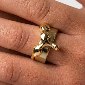 May include: A polished gold-tone ring featuring a wide, smooth band topped with an asymmetrical, raised molten-style design that extends across the surface. The metal has a high-shine, reflective finish with organic, flowing curves.