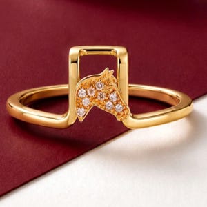 May include: A gold ring featuring a raised, rectangular bridge design. The center of the bridge is adorned with a horse head motif encrusted with small, clear, round-cut stones that catch the light, creating a sparkling, detailed appearance.