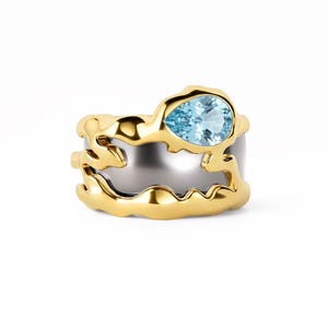 May include: A two-tone ring featuring a smooth silver-colored band encased within a textured, organic-shaped gold-colored frame. A single pear-shaped, light blue gemstone is set prominently at the top of the gold-colored metal structure.