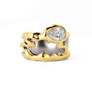 May include: A two-tone ring featuring a smooth silver-colored band encased by a textured, organic-shaped gold-colored frame. A single pear-cut clear gemstone is set within the gold frame, positioned at the top of the ring's irregular design.