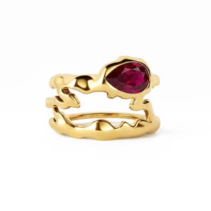 May include: A gold ring featuring an organic, molten-style band with an open, layered design. A single, pear-shaped, deep red gemstone is set prominently on the upper portion of the band, contrasting with the polished, reflective yellow gold surface.