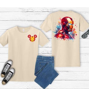 Puede incluir: Dos camisetas beige con dise&ntilde;os de Iron Man, una en la parte delantera y otra en la trasera. El dise&ntilde;o delantero es una peque&ntilde;a cara de Iron Man. El dise&ntilde;o trasero es una cabeza de Iron Man al estilo acuarela. La imagen tambi&eacute;n incluye vaqueros azules y zapatillas negras.