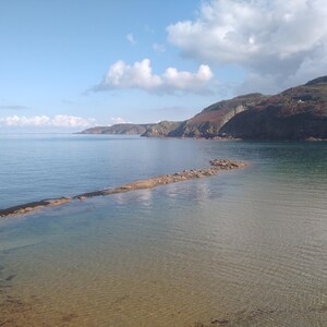 May include: A scenic coastal view featuring calm, clear water and a rocky breakwater extending into the sea. The sky is a bright blue with scattered white clouds. Hills covered in vegetation frame the water.