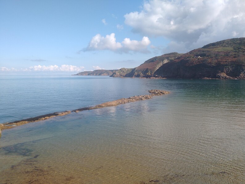 May include: A scenic coastal view featuring calm, clear water and a rocky breakwater extending into the sea. The sky is a bright blue with scattered white clouds. Hills covered in vegetation frame the water.