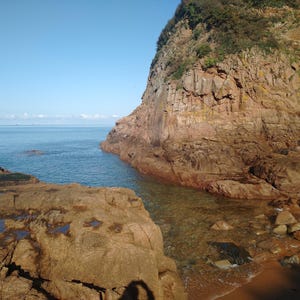 May include: A scenic coastal view featuring a rocky cliff face meeting the ocean. The water is clear and turquoise, with a sandy beach visible. The sky is a clear blue, creating a serene landscape.