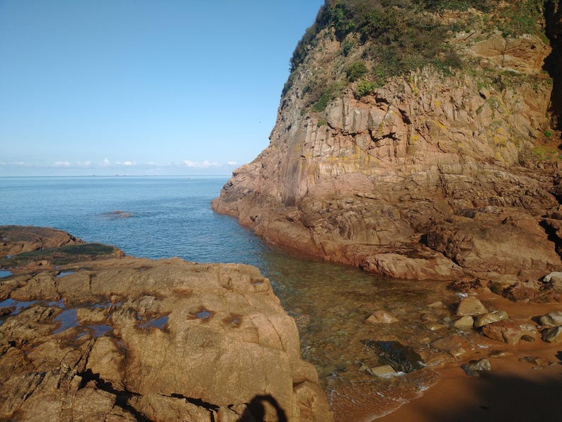 May include: A scenic coastal view featuring a rocky cliff face meeting the ocean. The water is clear and turquoise, with a sandy beach visible. The sky is a clear blue, creating a serene landscape.