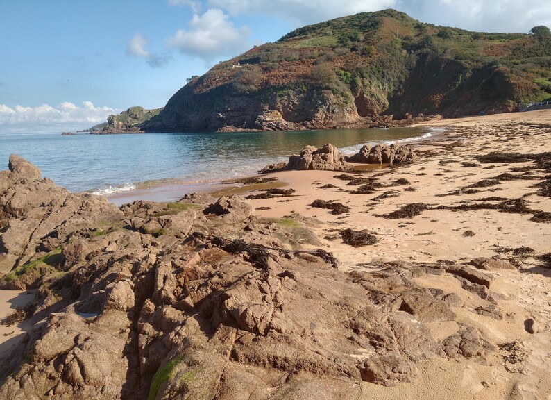 May include: A scenic coastal landscape featuring a sandy beach, rocky formations, and a calm ocean under a partly cloudy sky. A large hill covered in vegetation rises in the background, creating a natural, serene environment.