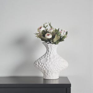 May include: A white ceramic vase with a textured surface, holding a bouquet of artificial flowers. The flowers are a mix of white and pink, with green leaves. The vase is on a dark surface.