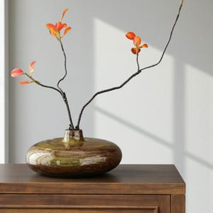 Op de afbeelding: Een ovale, decoratieve keramische vaas met een bruin en groen wervelpatroon, met takken met oranje en rode bladeren. De vaas staat op een houten oppervlak.