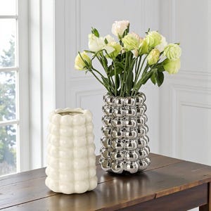 May include: Two decorative vases on a wooden table. One is a glossy, off-white color with a bubble design. The other is a silver vase with a similar bubble design, holding a bouquet of white and yellow flowers.