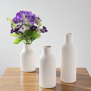 Puede incluir: Tres jarrones de cerámica blanquecina con detalles acanalados verticales. Un jarrón contiene un ramo de flores moradas y blancas con hojas verdes. Los jarrones están dispuestos sobre una superficie de madera.