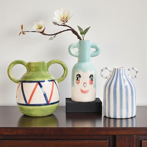Peut inclure: Trois vases décoratifs en céramique avec des anses. Le vase vert présente un motif blanc et rouge. Le vase bleu a un visage. Le vase rayé a des rayures verticales bleues et blanches. Une branche avec des fleurs blanches est dans le vase bleu.