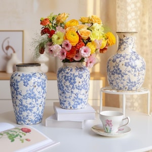 Könnte beinhalten: Drei Keramikvasen mit blauen Blumenmustern und ein Tee-Set auf einem weißen Tisch. Die Vasen sind mit bunten Blumen gefüllt. Die Tasse und Untertasse haben ein Blumenmuster.