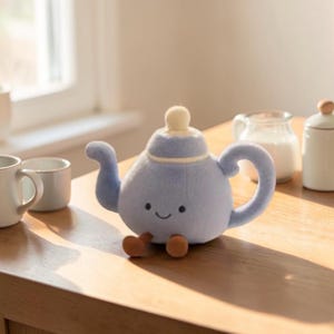 May include: A light blue plush teapot with a smiling face, a curved spout, and a rounded handle. The teapot has a small, round lid and two small brown feet. The teapot is sitting on a wooden surface.